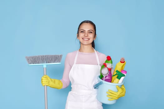 happy woman in yellow rubber gloves and cleaner apron holding bucket of detergents and broom on blue background.