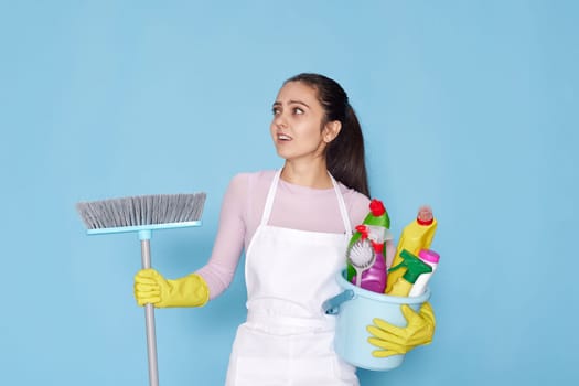 skeptic and nervous, frowning woman in gloves and cleaner apron holding bucket of detergents and broom on blue background.