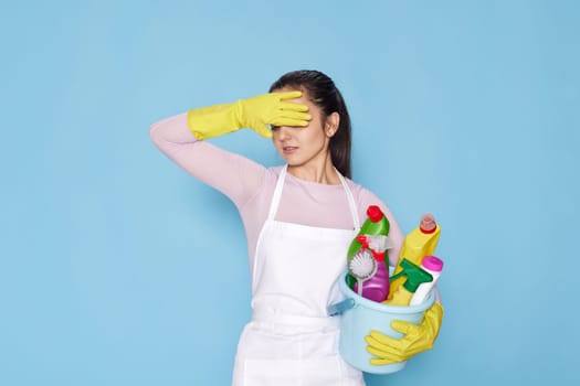 tired caucasian woman in gloves holding bucket with cleaning supplies and broom on blue background.