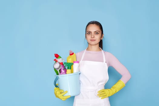 calm caucasian woman in gloves holding bucket with cleaning supplies isolated on blue background.