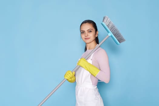 happy caucasian woman in rubber gloves and cleaner apron holding broom on blue background.