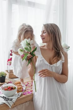 A little blonde girl with her mom on a kitchen countertop decorated with peonies. The concept of the relationship between mother and daughter. Spring atmosphere.