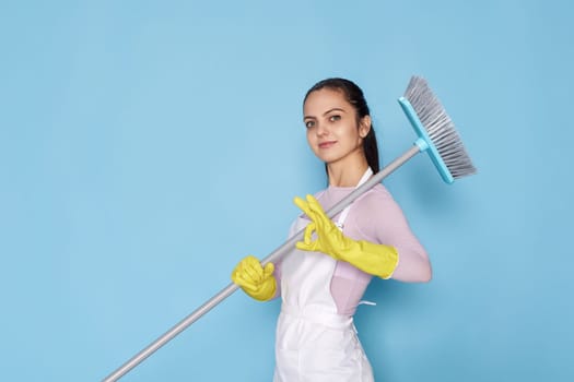 caucasian woman in rubber gloves and cleaner apron holding broom and showing ok gestureon blue background.
