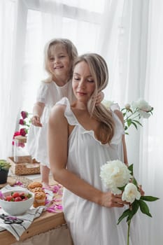 A little blonde girl with her mom on a kitchen countertop decorated with peonies. The concept of the relationship between mother and daughter. Spring atmosphere.