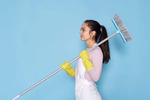 caucasian woman in rubber gloves and cleaner apron holding broom on blue background.