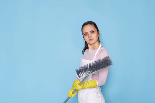 nervous, frowning woman in rubber gloves and cleaner apron with broom on blue background.