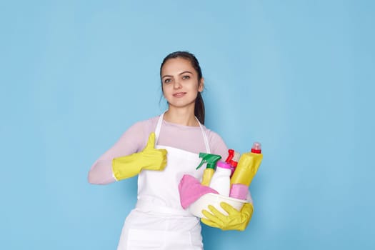 happy caucasian woman in gloves and cleaner apron holding bucket of detergents and showing Ok gesture on blue background.