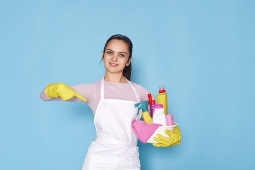 caucasian woman in rubber gloves pointing fingers at bucket with cleaning supplies on blue background.