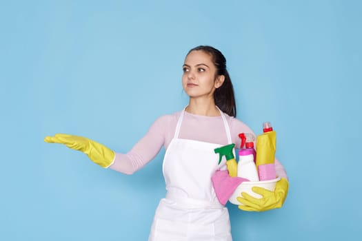 caucasian woman in rubber gloves with bucket with cleaning supplies holding copy space on her palm on blue background.