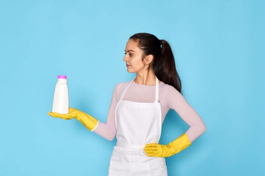 beautiful caucasian woman in yellow rubber gloves and cleaner apron holding bottle of bleach on blue background.