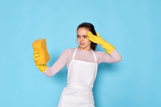 dissatisfied woman in yellow rubber gloves and cleaner apron holding sponge on blue background.