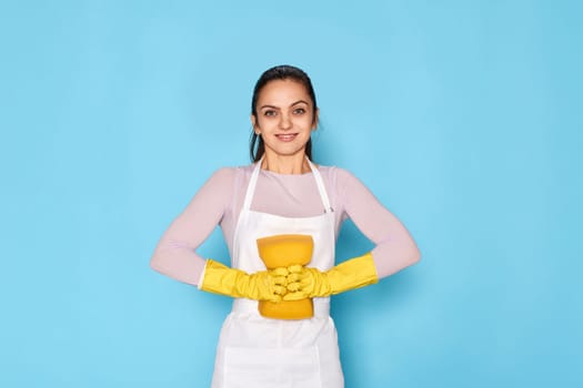 happy woman in yellow rubber gloves and cleaner apron holding sponge on blue background.