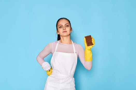 diligent woman in rubber gloves and cleaner apron cleaning with sponge on blue background.