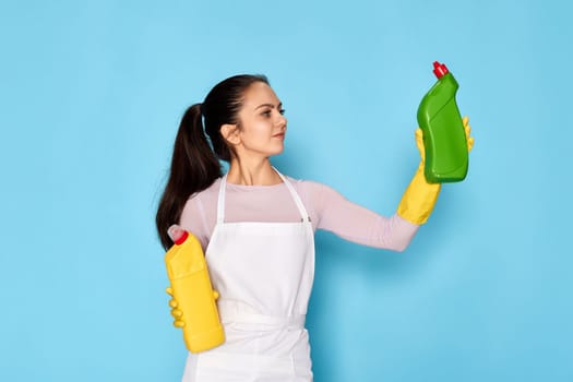 beautiful woman in rubber gloves and cleaner apron chooses between two cleaning products on blue background.
