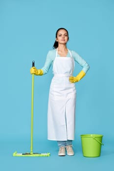 professional cleaner girl in rubber gloves and cleaner apron washing floor with mop on blue background. Full length