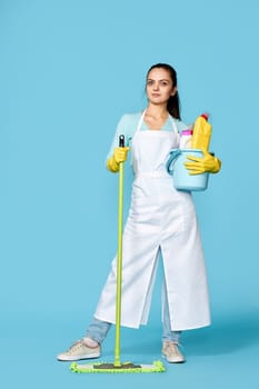 pretty woman in rubber gloves and cleaner apron with cleaning supplies and mop on blue background