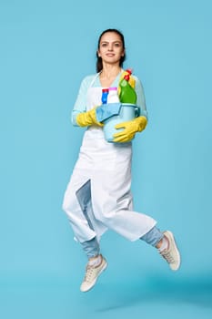 happy cheerful woman in rubber gloves and cleaner apron with cleaning supplies jumping on blue background. Full length