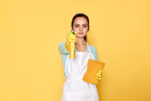 caucasian woman in yellow rubber gloves and cleaner apron with sponge and detergent sprayer on yellow background.