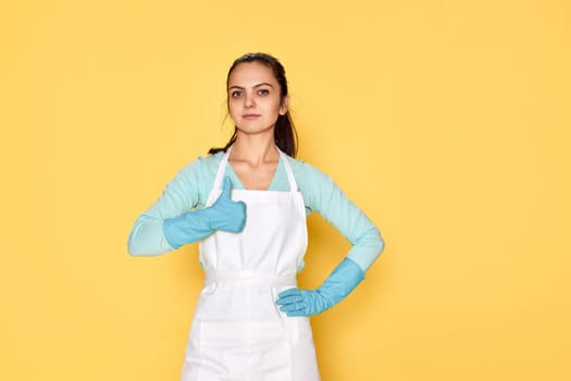happy caucasian woman in blue rubber gloves and cleaner apron showing thumbs up gesture on yellow background. cleaning