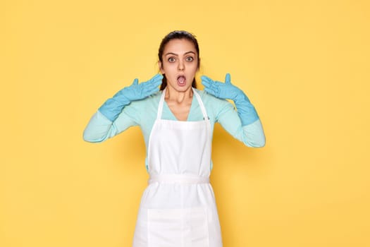 surprised young woman in blue rubber gloves and cleaner apron on yellow background.