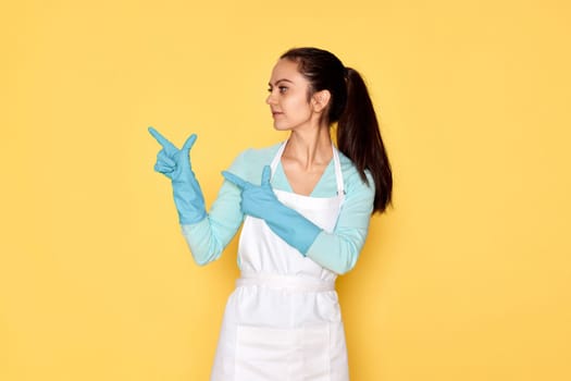 beautiful caucasian woman in rubber gloves pointing fingers aside at copy space on yellow background. cleaning
