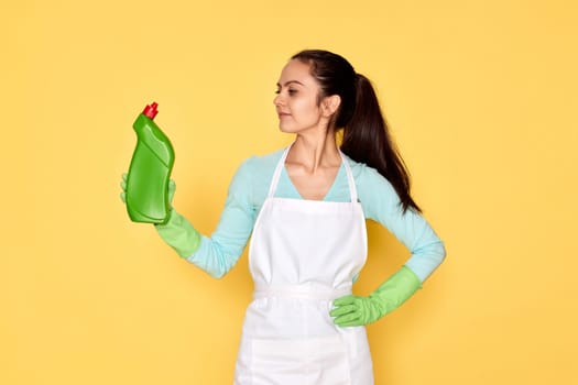 beautiful caucasian woman in rubber gloves and cleaner apron holding bottle of detergent on yellow background.