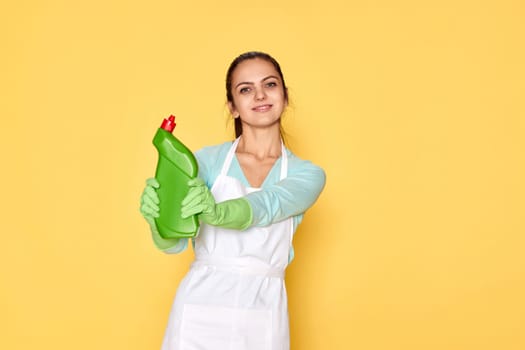 beautiful caucasian woman in rubber gloves and cleaner apron holding bottle of detergent on yellow background.