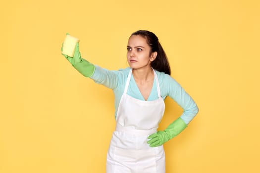 young confident woman in rubber gloves and cleaner apron cleaning with yellow sponge on yellow background.