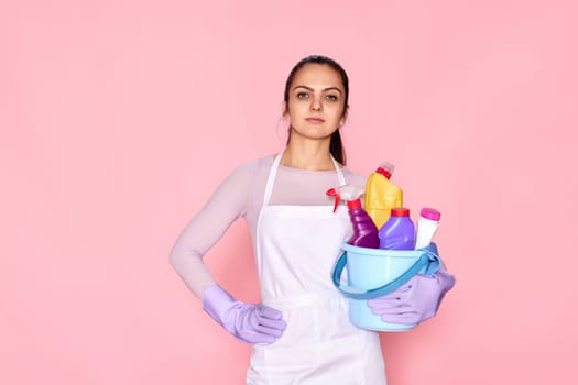 pretty caucasian woman in gloves holding bucket with cleaning supplies isolated on pink background.