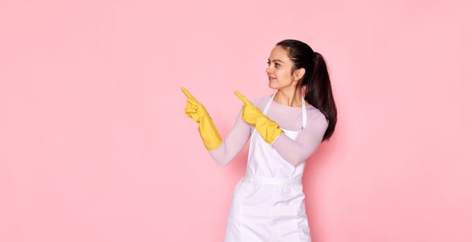 beautiful caucasian woman in rubber gloves pointing fingers aside at copy space on pink background. cleaning