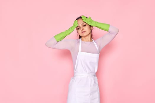 tired woman in green gloves and cleaner apron on pink background