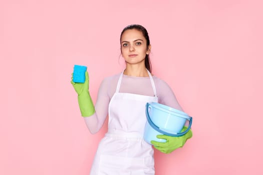 young confident woman in rubber gloves and cleaner apron cleaning with blue sponge and bucket on pink background.