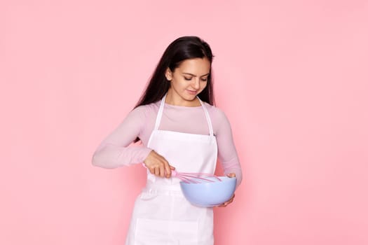Pretty cooker woman holding whisk and bowl on pink background