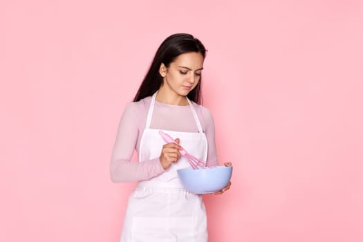 Pretty housekeeper baker woman holding whisk and bowl on pink background