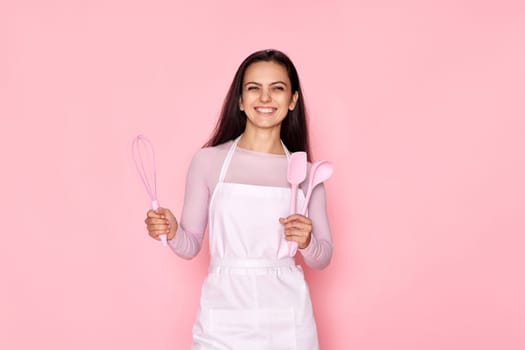 happy cooker woman in chef apron holding whisk, spoon and spatula on pink background