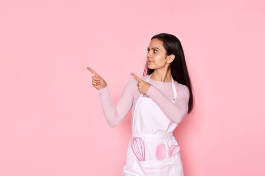 pretty woman in chef apron in whose pocket a whisk and a spatula pointing fingers aside at copy space on pink background