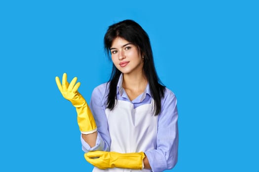 pretty woman in yellow rubber gloves and cleaner apron looking at camera isolated on blue background. cleaning