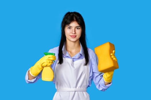 caucasian woman in yellow rubber gloves and cleaner apron with sponge and detergent sprayer isolated on blue background. cleaning