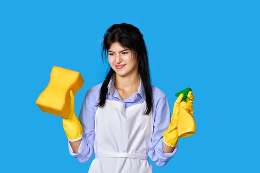 nervous, frowning caucasian woman in yellow rubber gloves and cleaner apron with sponge and detergent sprayer isolated on blue background. cleaning