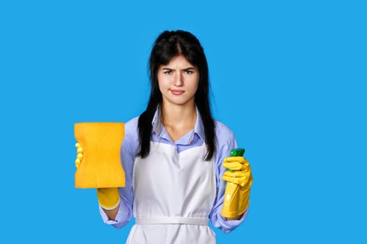 unhappy upset caucasian woman in yellow rubber gloves and cleaner apron with sponge and detergent sprayer isolated on blue background. cleaning