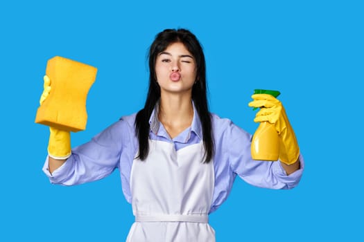 caucasian woman in yellow rubber gloves and cleaner apron with sponge and detergent sprayer isolated on blue background. cleaning