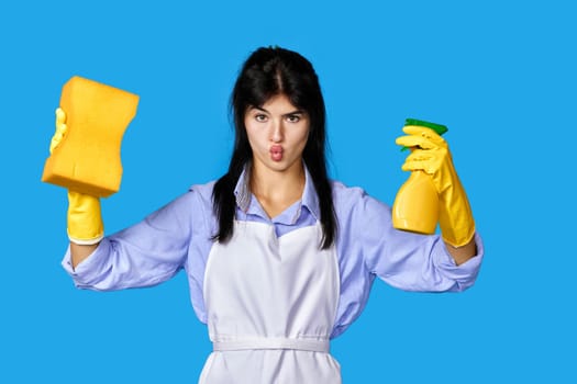 caucasian woman in yellow rubber gloves and cleaner apron with sponge and detergent sprayer isolated on blue background. cleaning