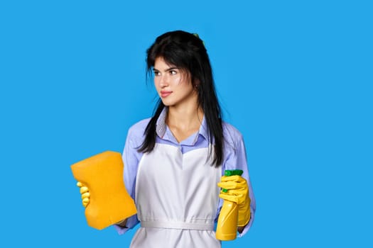upset caucasian woman in yellow rubber gloves and cleaner apron with sponge and detergent sprayer isolated on blue background. cleaning