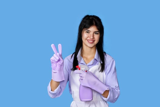 smiling woman in rubber gloves and cleaner apron showing victory gesture isolated on blue background. cleaning