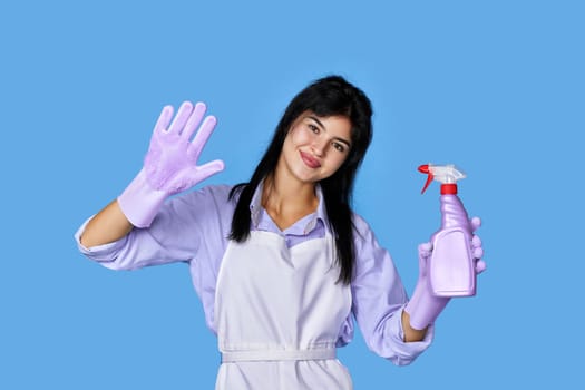 happy woman in purple rubber gloves and cleaner apron with detergent sprayer isolated on blue background. cleaning