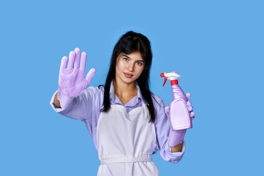 beautiful woman in rubber gloves and cleaner apron showing stop gesture isolated on blue background. focus on glove
