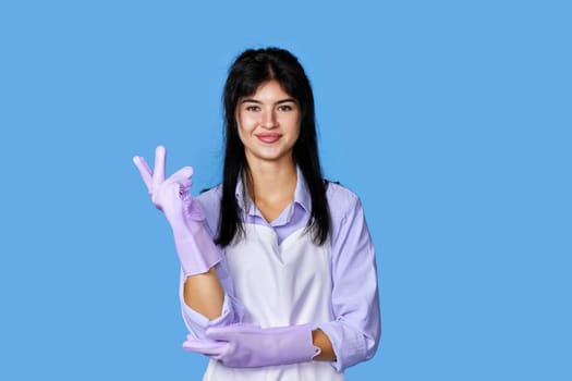 pretty woman in rubber gloves and cleaner apron looking at camera isolated on blue background. cleaning