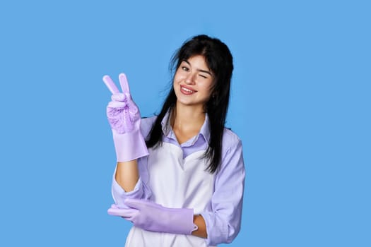 cheerful woman in rubber gloves and cleaner apron winking looking at the camera and showing victory gesture isolated on blue background. cleaning