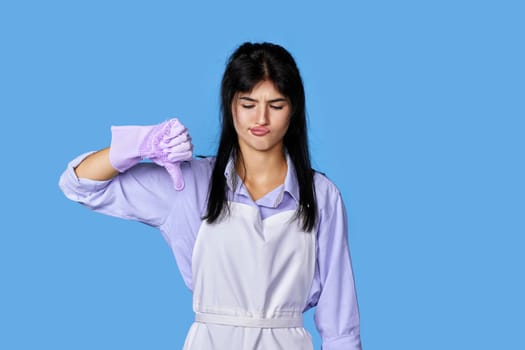 displeased woman in violet rubber gloves and cleaner apron showing thumbs down sign isolated on blue background. cleaning