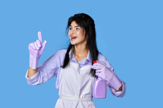 beautiful woman in violet rubber gloves and apron with detergent sprayer holding index finger up with new idea isolated on blue background. cleaning